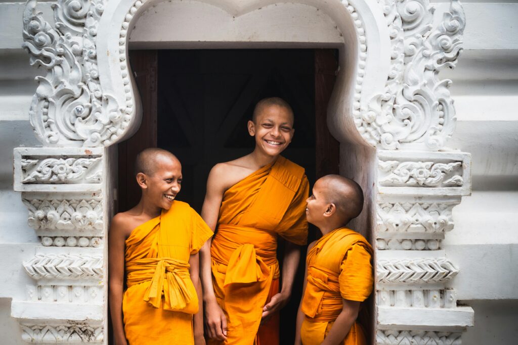 Three young Buddhist monks in saffron robes smiling in a decorative doorway in India.